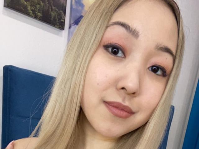 RenyLime has blonde hair, light skin, and brown eyes with pink eyeshadow. She's wearing nude lipstick, and has a small mole on her right cheek. Blue chair in background.