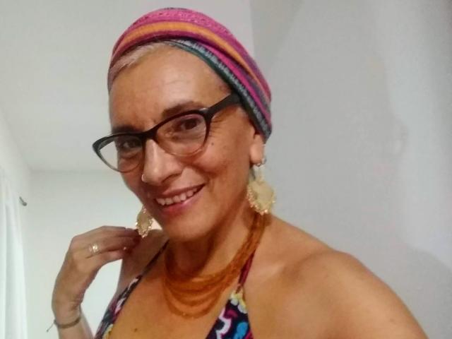 EstelaMarisi with gray hair, black glasses, pink and purple striped headscarf, colorful halter top, orange necklace, and large lace earrings, smiling, showing her tits.