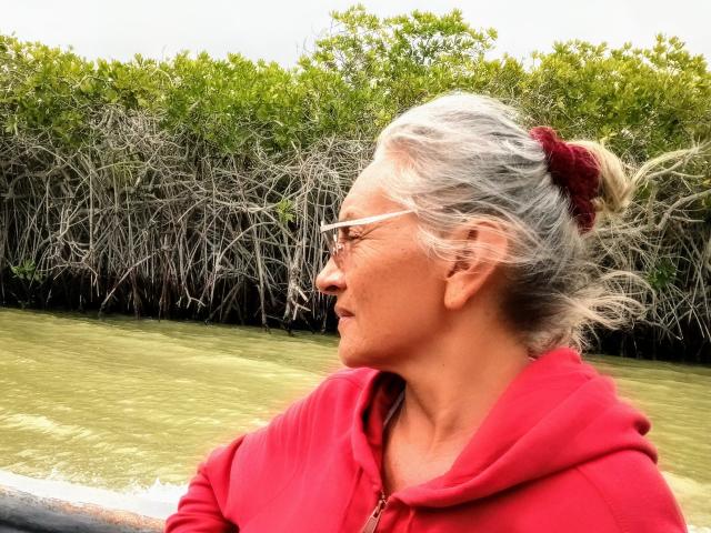 EstelaMarisi with gray hair in a red scrunchie, wearing a red hoodie and white glasses, looking to the side, by a greenish waterway with a dense, leafy backdrop.
