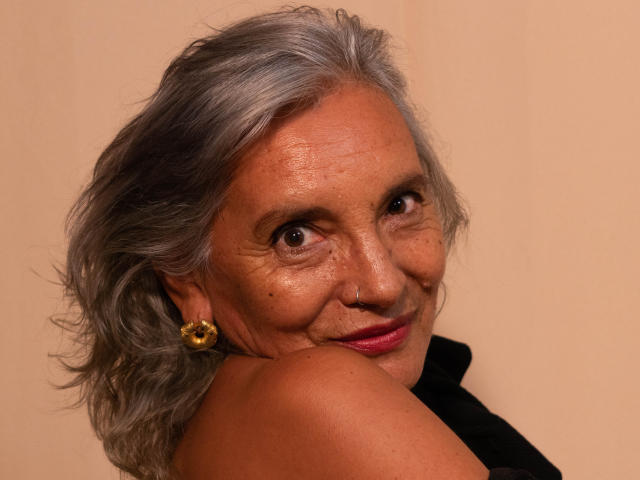 EstelaMarisi has gray, wavy hair, brown eyes, and is smiling with pink lipstick. She's wearing yellow earrings and has her shoulder up, showing a tan, wrinkled face.