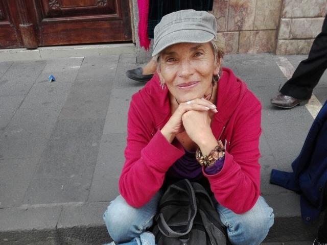 EstelaMarisi, elderly woman, crouching on sidewalk, wearing red sweater, gray cap, blue jeans, purple shirt, bracelet, black bag, smiling, hands resting on chin, grey hair, wrinkled face.