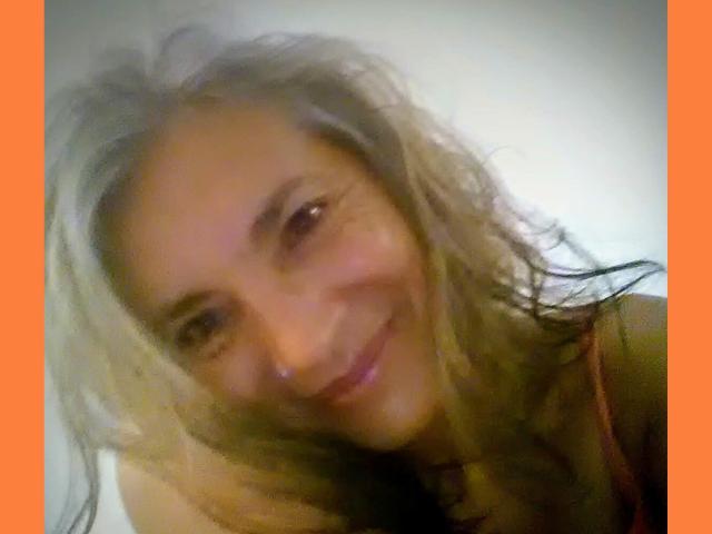Blurry selfie of EstelaMarisi with messy blonde hair, smiling slightly, showing a small nose and brown eyes, wearing a red top, against a white background.