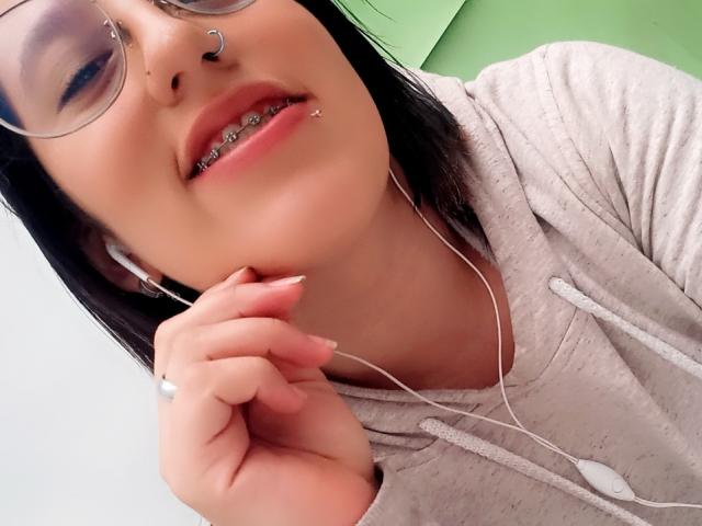 NatalieHarris with glasses, nose ring, and lip piercing, wearing a grey hoodie, holding white earbuds, showing metal teeth, and black hair, slightly smiling.