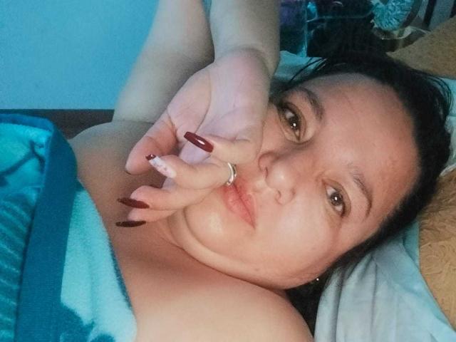 SkayLaraWalker tiene ojos marrones, cabello oscuro y uñas rojas que cubren su boca, y está acostada en una cama con sábanas azules y blancas, mostrando sus hombros desnudos y un anillo..