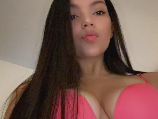 IssaPuasini with long black hair, pink bra, and pouty lips, showing her big, round, perky tits. Her skin is light, and she's looking at the camera.