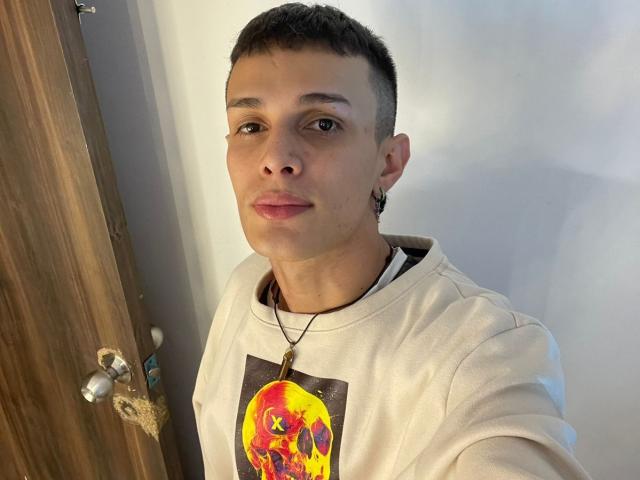 SlimLewis, short brown hair, brown eyes, white shirt with colorful graphic, necklace with cross, small earring, wooden door, white wall, slight smile, casual selfie.