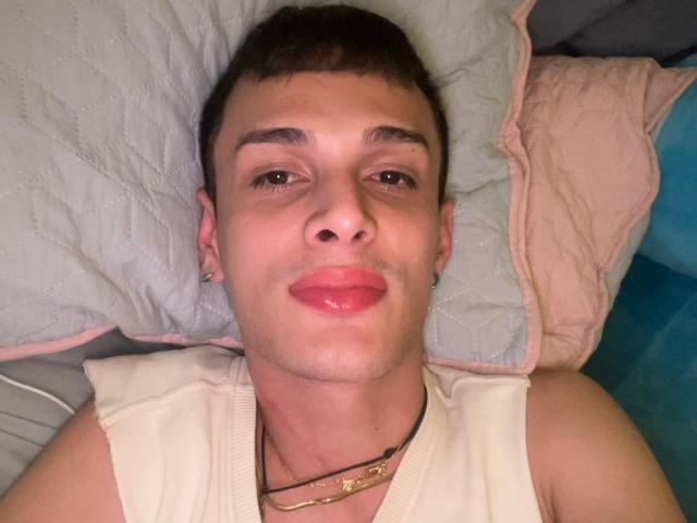 SlimLewis with short brown hair, light skin, and glossy pink lips, wearing a white sleeveless shirt, lying on a pillow, looking at the camera with a slight smile.