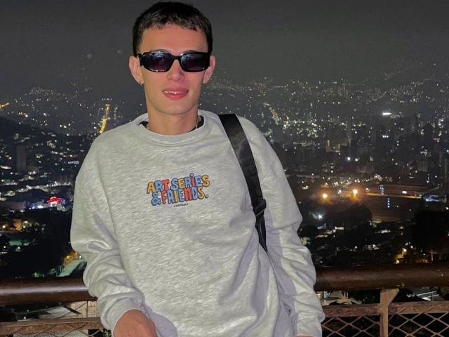 SlimLewis with short brown hair, wearing black sunglasses and a gray "Angels" sweatshirt, standing on a balcony at night with a city skyline in the background.