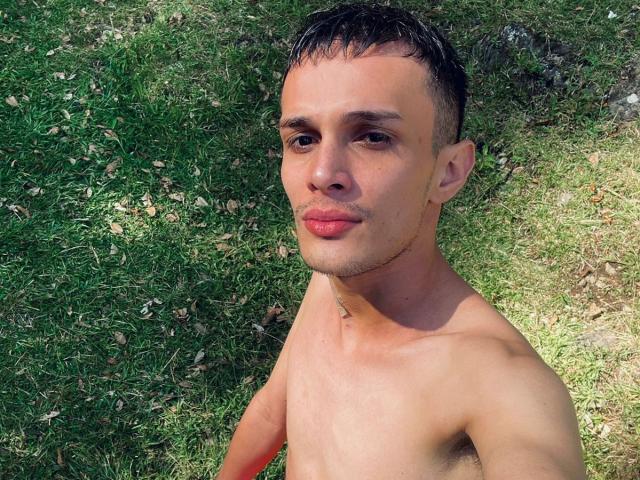 SlimLewis, shirtless with short dark hair, light skin, and a slight smile, lying on green grass. His small, slightly pointed ass and slim chest are visible.