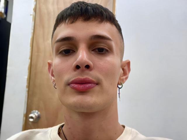 SlimLewis has short brown hair, a light skin tone, and wears a small black earring in his right ear with pinkish lipstick. He's looking straight at the camera.