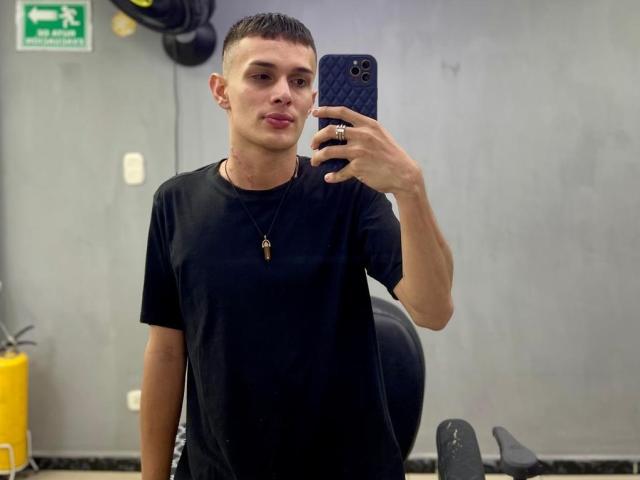 SlimLewis with short black hair, wearing a black tee and necklace, taking a selfie with a diamond-patterned phone in a gray-walled room. His face is slightly pouty.