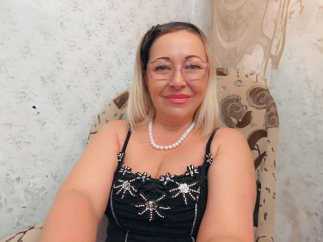 LoriExcite tem cabelo loiro, usa óculos e um colar de pérolas. Ela está vestindo um vestido preto com padrões brilhantes, está sorrindo e tem uma bunda charmosa e curvilínea..