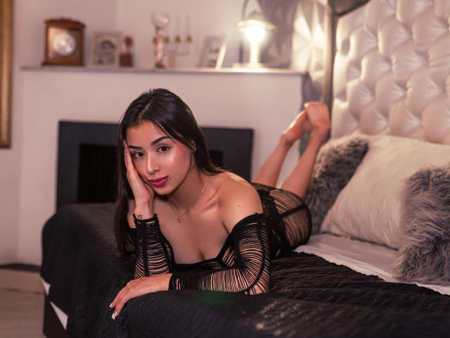 AntonellaaRossy lies on a bed, wearing a black, off-shoulder, striped dress, her tits slightly visible, head on hand, legs up, in a cozy, softly lit bedroom.
