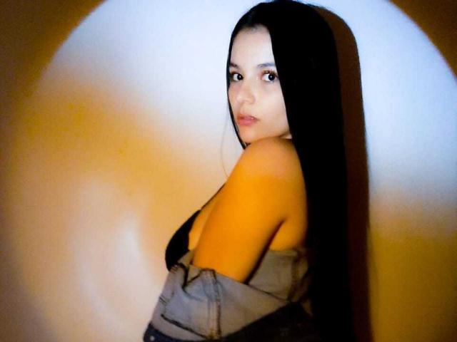 HannaSpencer with long black hair, looking over her shoulder, wearing a strapless top, exposing her bare back and small tits. Soft yellow light highlights her face and upper body.