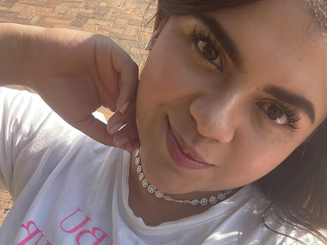 MissSofiia heeft een medium bruine huid, donkerbruine ogen en een schattige glimlach. Ze draagt een witte blouse, een diamanten ketting en heeft haar hand in de buurt van haar gezicht..