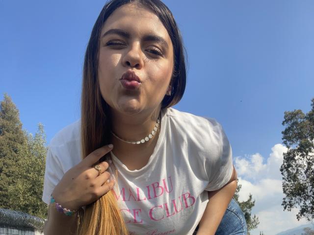 MissSofiia, con labios fruncidos, cabello castaño, camisa blanca, collar y anillo, mirando hacia abajo a la cámara, al aire libre en un día soleado. Su trasero es ligeramente visible, inclinándose hacia adelante..