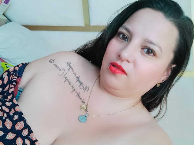 SkayLaraWalker with red lips, black hair, and a tattoo on her left shoulder, wearing a patterned dress, looking at the camera. Her tits are slightly visible.