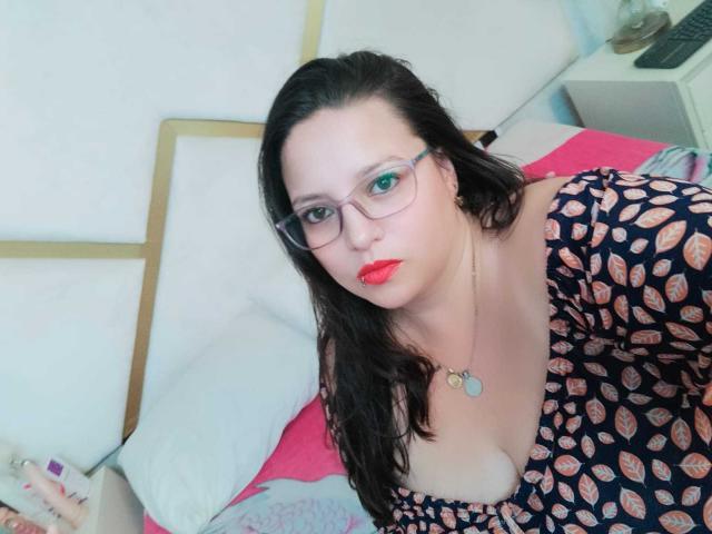 SkayLaraWalker, usando óculos, batom vermelho e com cabelo ondulado escuro, vestindo um vestido estampado com decote, sentada em uma cama, olhando para a câmera..