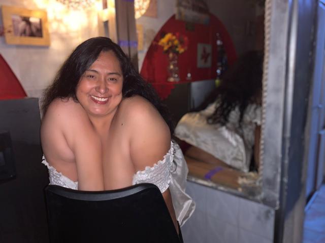 TistMature, una mujer latina de curvas, con cabello largo y oscuro, sonríe mientras se inclina hacia adelante en una silla, con los hombros desnudos y la parte superior de los senos visibles, vistiendo una blusa blanca con un hombro descubierto..