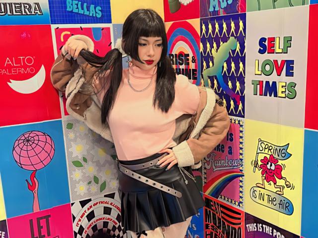 SabriPrettty with black pigtails, pink turtleneck, brown jacket, black leather skirt, holding hair, wearing a silver necklace, standing in front of colorful, patterned posters.