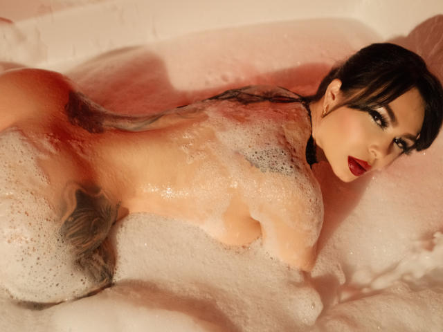 GabrielaAroms, nue, dans une baignoire avec de la mousse rose, allongée sur le ventre, montrant ses fesses, son dos et son rouge à lèvres rouge, regardant l'appareil photo avec ses cheveux noirs et ondulés..