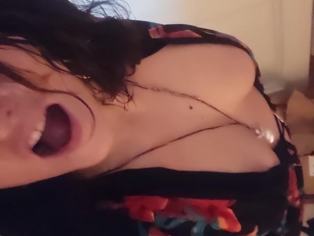 Cellaa69 tem cabelos pretos e molhados, boca aberta e seios grandes, vestindo uma blusa preta com decote baixo e estampas florais vermelhas, mostrando um colar entre seus seios..