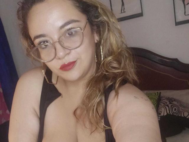 NaugthyCurvy, com cabelo loiro ondulado, óculos, batom vermelho, brincos de argola dourados e uma regata preta, mostrando seus seios, em uma selfie no quarto..