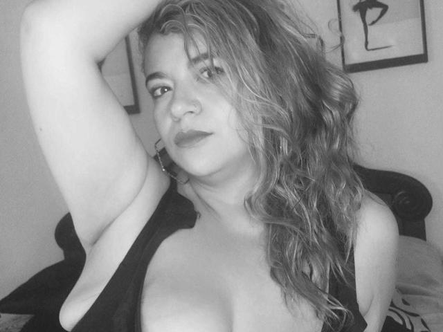 Zwart-wit foto van NaugthyCurvy met golvend, lichtbruin haar, die een arm omhoog steekt, met een decolleté in een top met lage hals, en die in de camera kijkt met een licht geopende mond..