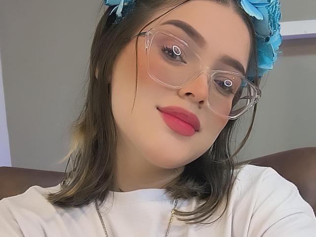 Kim: Sommer mit klaren Brillen, einem blauen Blumenhaarreif, pinkem Lippenstift, einem weißen Hemd und braunen, schulterlangen Haaren. Ihr Gesicht ist leicht geneigt, und sie schaut direkt in die Kamera..
