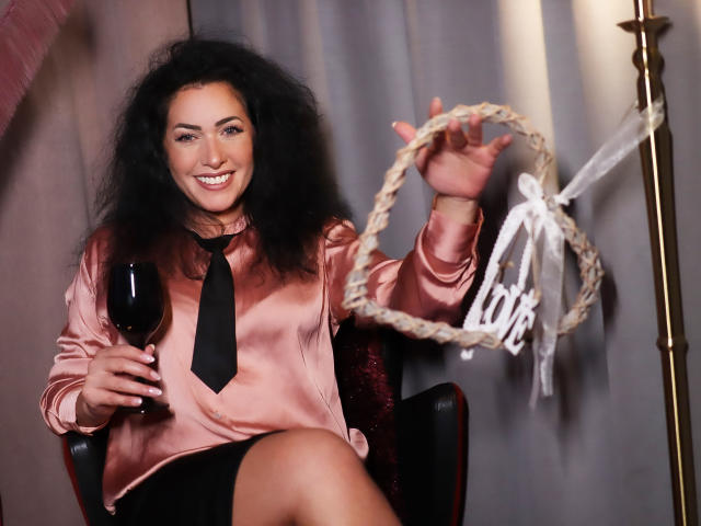 AnelisaReys, avec ses cheveux noirs bouclés, souriante, portant une chemise rose en satin et une cravate noire, tenant un verre de vin rouge, et levant un anneau tressé avec un ruban blanc..