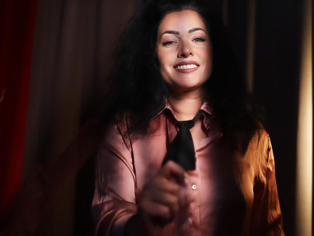 AnelisaReys, avec ses cheveux noirs bouclés, souriante, portant une chemise rose brillante et une cravate noire, tenant un objet noir devant elle. Ses seins sont partiellement visibles..