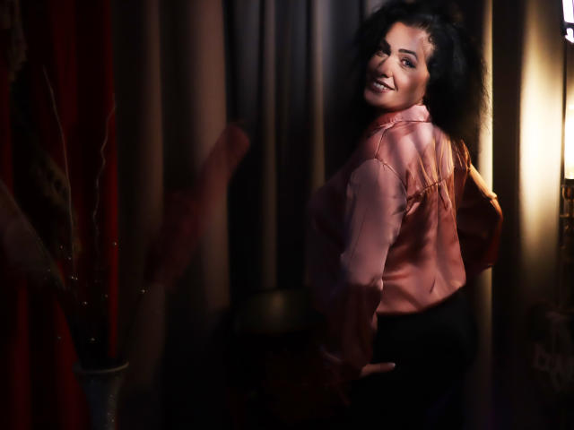 AnelisaReys, met krullend zwart haar, lachend, gekleed in een roze satijnhemd, zwarte broek, en staand met een hand op haar achterwerk, kijkt over haar schouder in een slecht verlichte kamer..