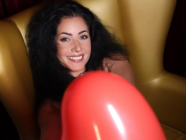 AnelisaReys, avec ses cheveux noirs bouclés, sourit en regardant un ballon rouge, assise sur une chaise jaune. Ses seins sont partiellement visibles, et elle a un air mignon et séduisant..