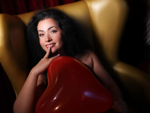 Anelisa, met krullend zwart haar, lichte huid en een schattige glimlach, houdt een rood, glanzend object in haar schoot, zittend in een gele stoel, met haar vinger tegen haar lippen..