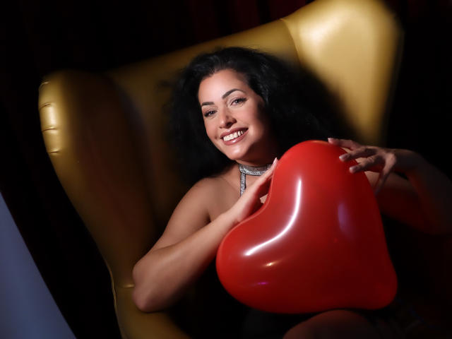 AnelisaReys, com cabelos pretos encaracolados, sorrindo, segurando um balão vermelho brilhante em formato de coração, sentada em uma cadeira amarela, usando um colar de prata, fundo escuro..