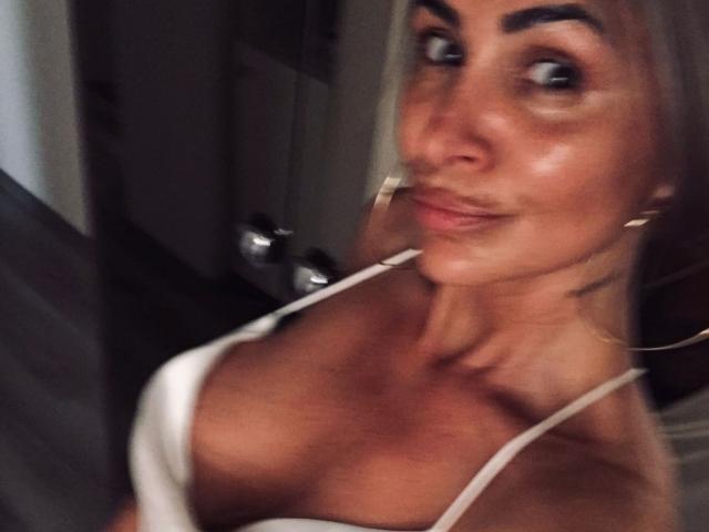 BriliantOne with tan skin and brown eyes, smiling slightly, wearing a white strap dress, showing her tits, in a blurry selfie, with subtle makeup and a gentle smile.