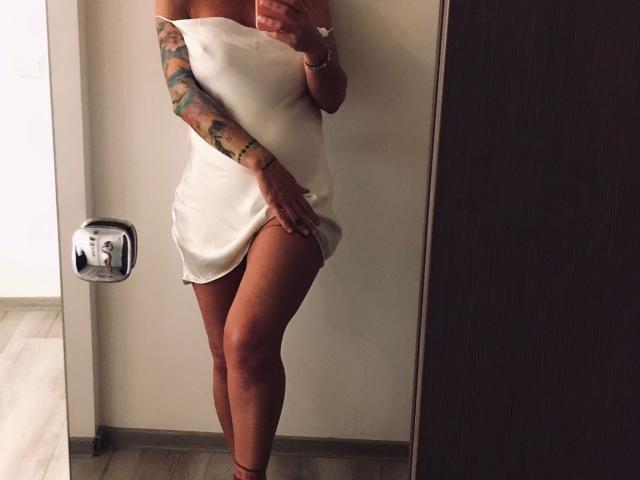 BriliantOne in a white dress, holding her crotch, showing her ass, tattooed arm, and phone in hand, standing in a dimly lit room with a mirror.