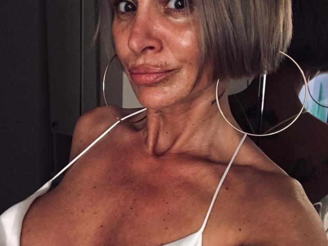 BriliantOne with short gray bob, large hoop earrings, and white halter top, showing tan skin and small breasts, smiling slightly. Her lips are naturally pink, and she has tattoos.