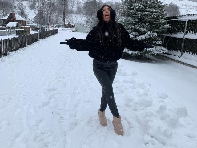 CataleyaLee, snow-covered street, black hoodie, green pants, beige boots, black gloves, snowflakes, open mouth, arms out, mid-step, winter, snow, wooden fence, trees, house in background.
