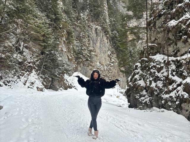 CataleyaLee stands in a snowy forest, arms spread, wearing a black hoodie and gray pants, smiling with snow on her face and boots in the white path.