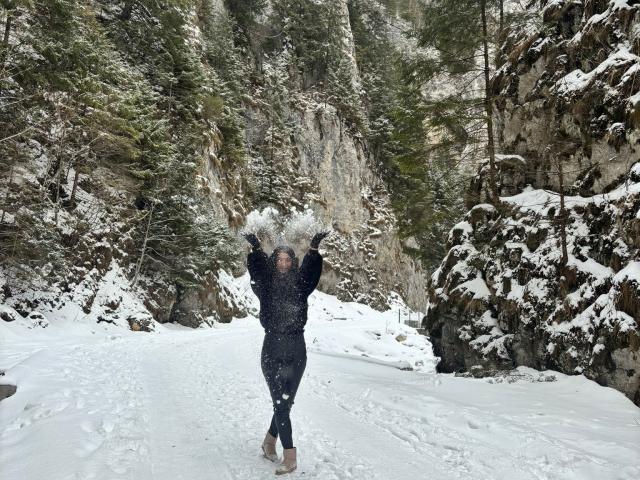 CataleyaLee is standing in a snowy forest, arms raised, wearing a black coat with fur trim, snow falling on her, and a snow-covered path in front of her.