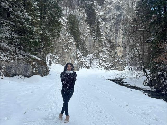 CataleyaLee stands in a snowy forest, snowflakes on her black coat, green pants, and beige boots, with a stream to the right and trees around.