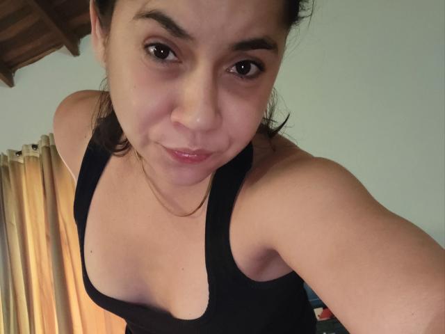 MsJustineBeaux with medium skin, dark eyes, and small tits in a black tank top, smiling slightly, brown hair in a messy bun, wearing a thin gold necklace, in a selfie.