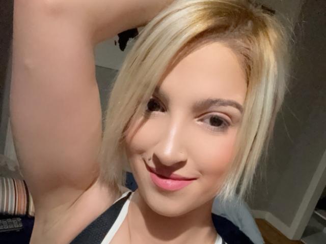Ajunas blonde hair, fair skin, smiling face, pink lips, raised arm, nose ring, dark eyebrows, white top, soft lighting, casual selfie, slight pink blush on cheeks.