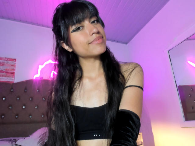 SalomAckay met lang zwart haar, gekleed in een zwart topje en mouwloze fluwelen mouwen, met een lichte glimlach, roze neonlichten op de achtergrond, een paars plafond, een spiegel aan de rechterkant..