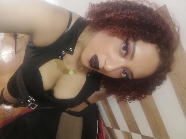 VioletaSwitch has curly red hair, dark lipstick, and wears a black top with a deep cleavage, showing her tits. She's also got a gold necklace and black straps.