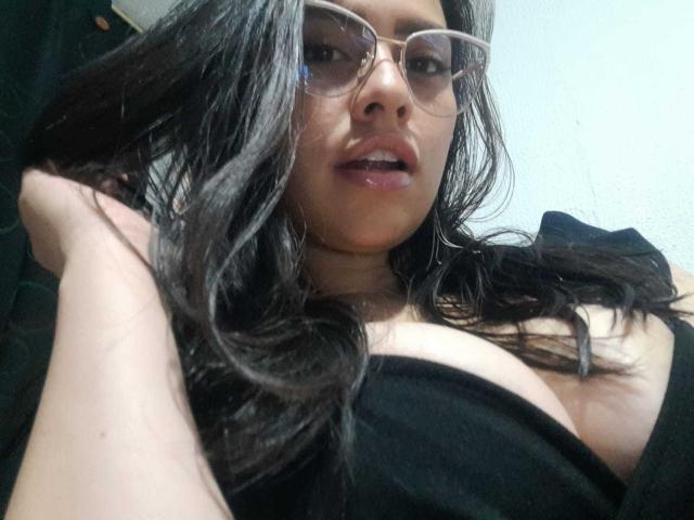 BellaSmithX69 with glasses, dark wavy hair, black strapless top, exposing large boobs, right hand in hair, slightly open mouth, light skin, looking at camera, indoors.