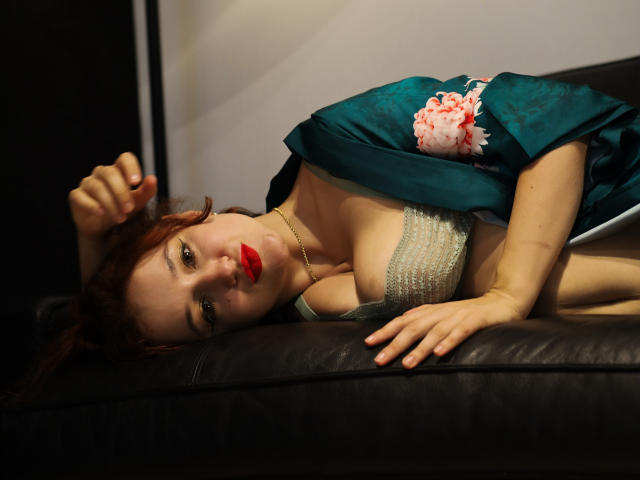 LailaNoire, with red lipstick, lies on a black leather couch, wearing a green robe with pink floral design, exposing her tits in a white lace bra.