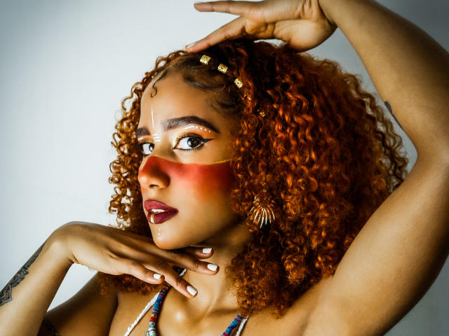Cinthia Walker mit lockigem, rot-orangefarbenem Haar, dunklem Lippenstift und auffälligem Augen-Make-up, die ihr Kinn hält und einen Arm hebt, trägt eine perlenbesetzte Halskette und kunstvolle Ohrringe und zeigt ein Tattoo..