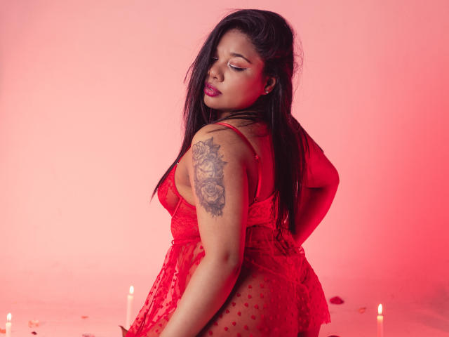 TriniShely with red lace lingerie, showing her ass and tattoo on her left arm, standing with candles below, her eyes closed and lips slightly parted. Red lighting.
